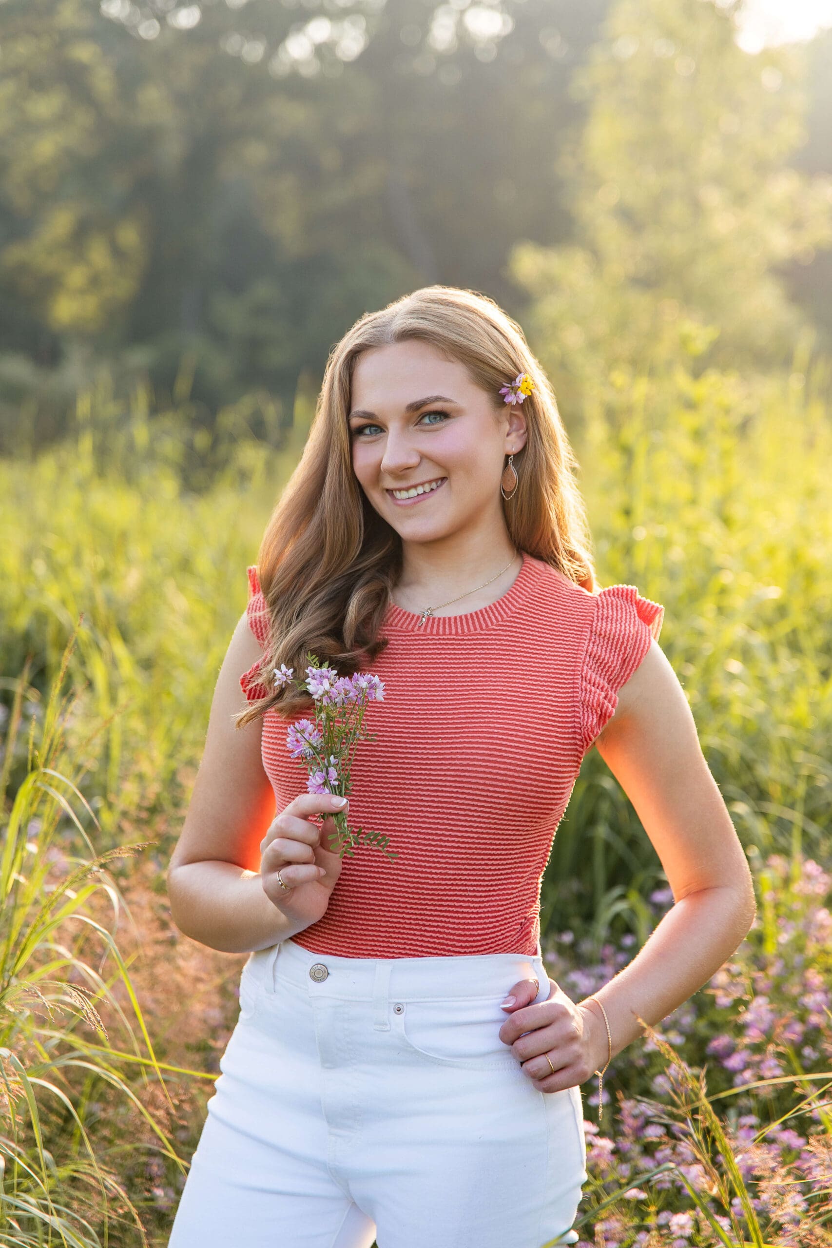 A happy high school senior in a red blouse holds wildflowers in a field at sunset