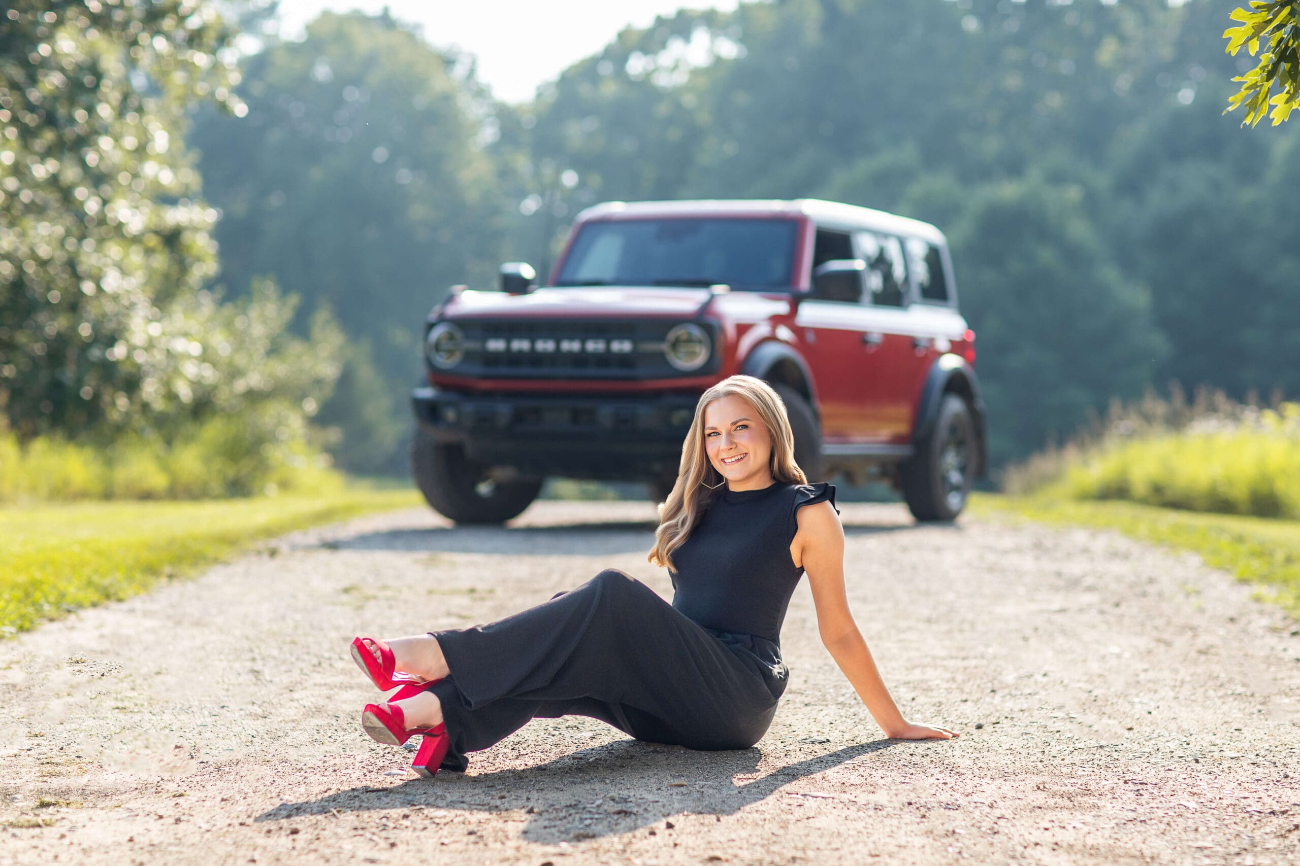 A smiling high school senior sits in a gravel road in front of her red truck in a black outfit after visiting boutiques in metro detroit