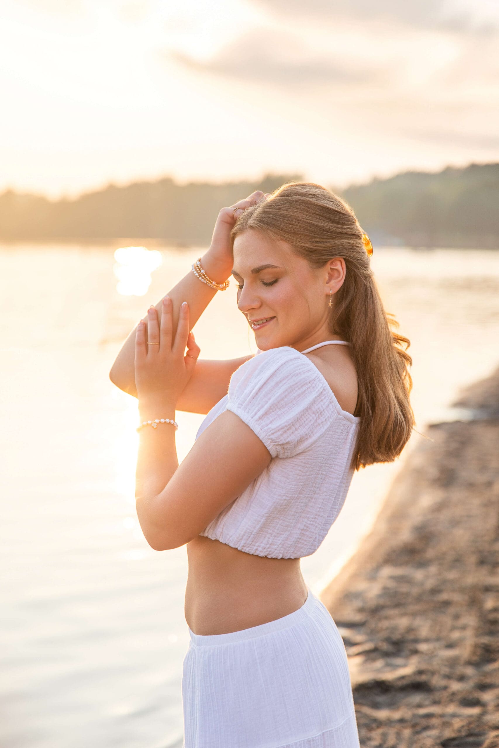 A high school senior in white. dances on the beach at sunset with a hand in her hair after visiting boutiques in metro detroit