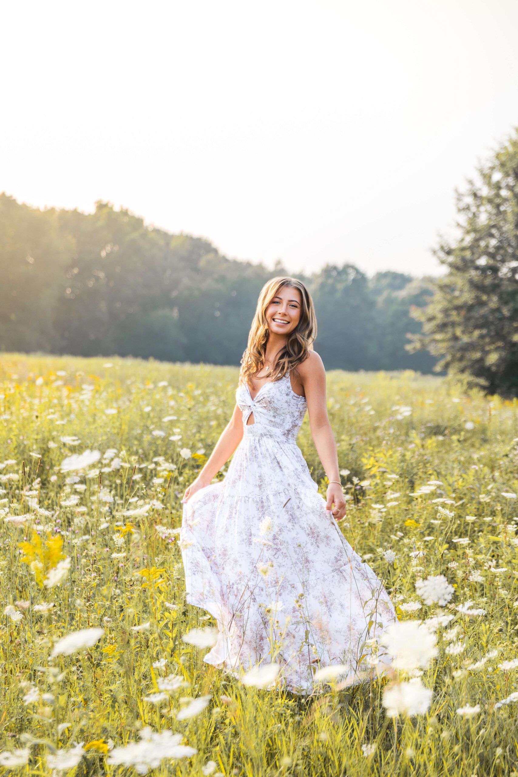 A high school senior smiles big while walking in a field of wildflowers at sunset in a flower print dress