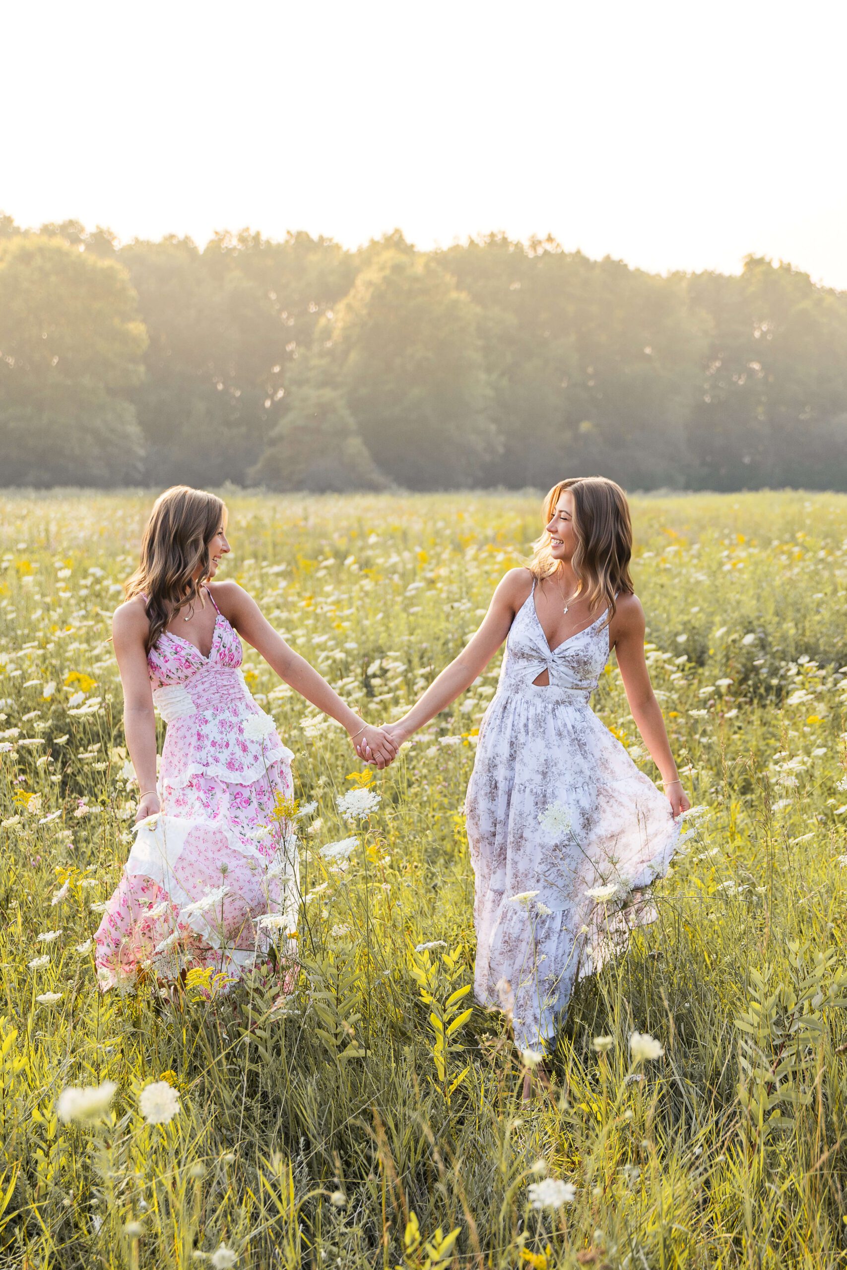 Sisters in pink and blue dresses walk ina. field of wildflowers holding hands at sunset