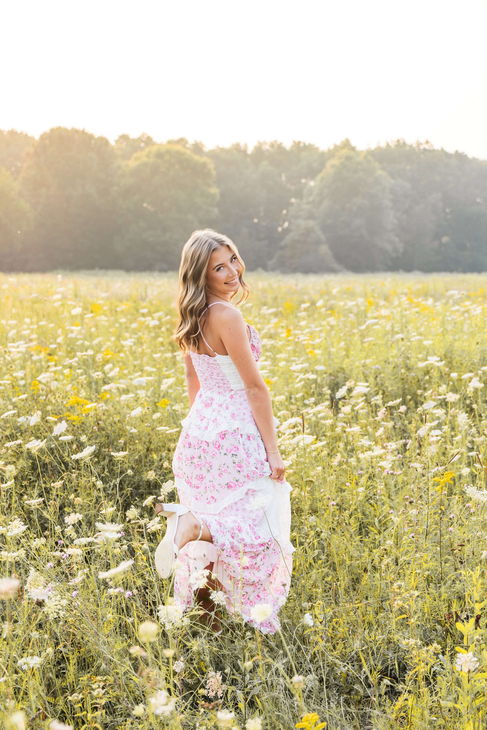 A smiling high school senior in a pink dress plays in a field of wildflowers at sunset while smiling over her shoulder after exploring detroit party venues