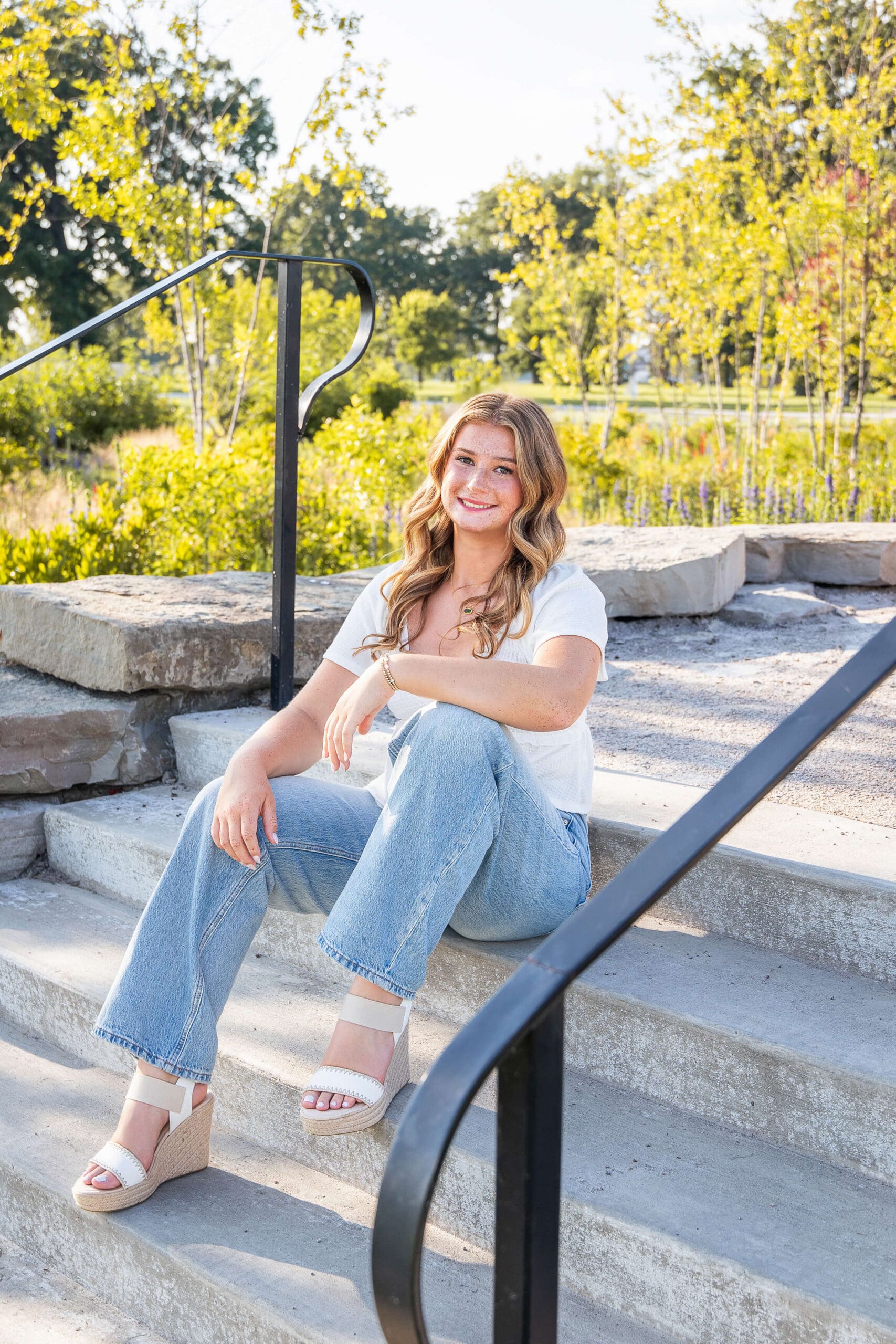 A high school grad in a white shirt and jeans smiles while sitting on garden steps at sunset