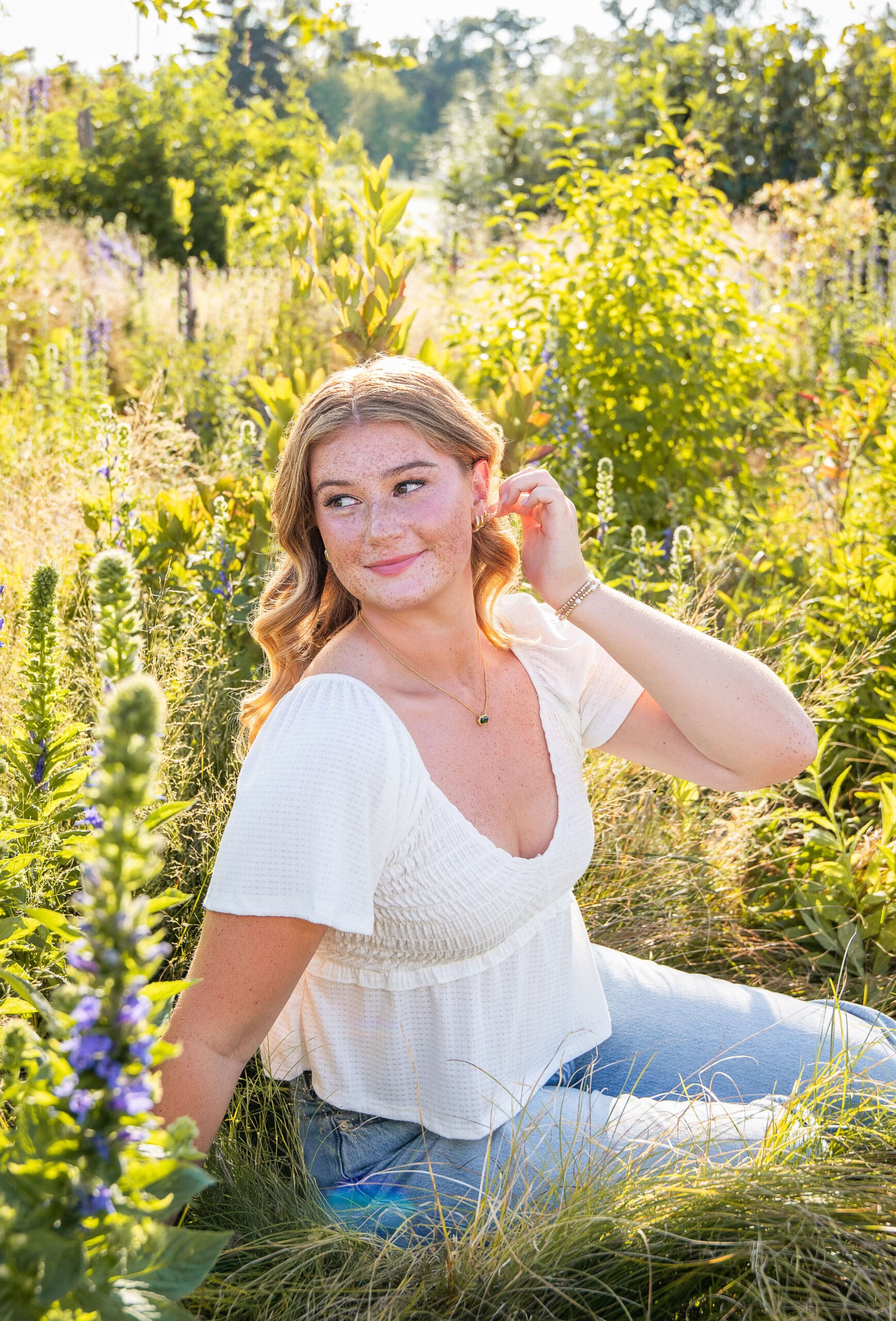A happy high school senior smiles over her shoulder while sitting in tall grasses with wildflowers after visiting hair salons in metro detroit
