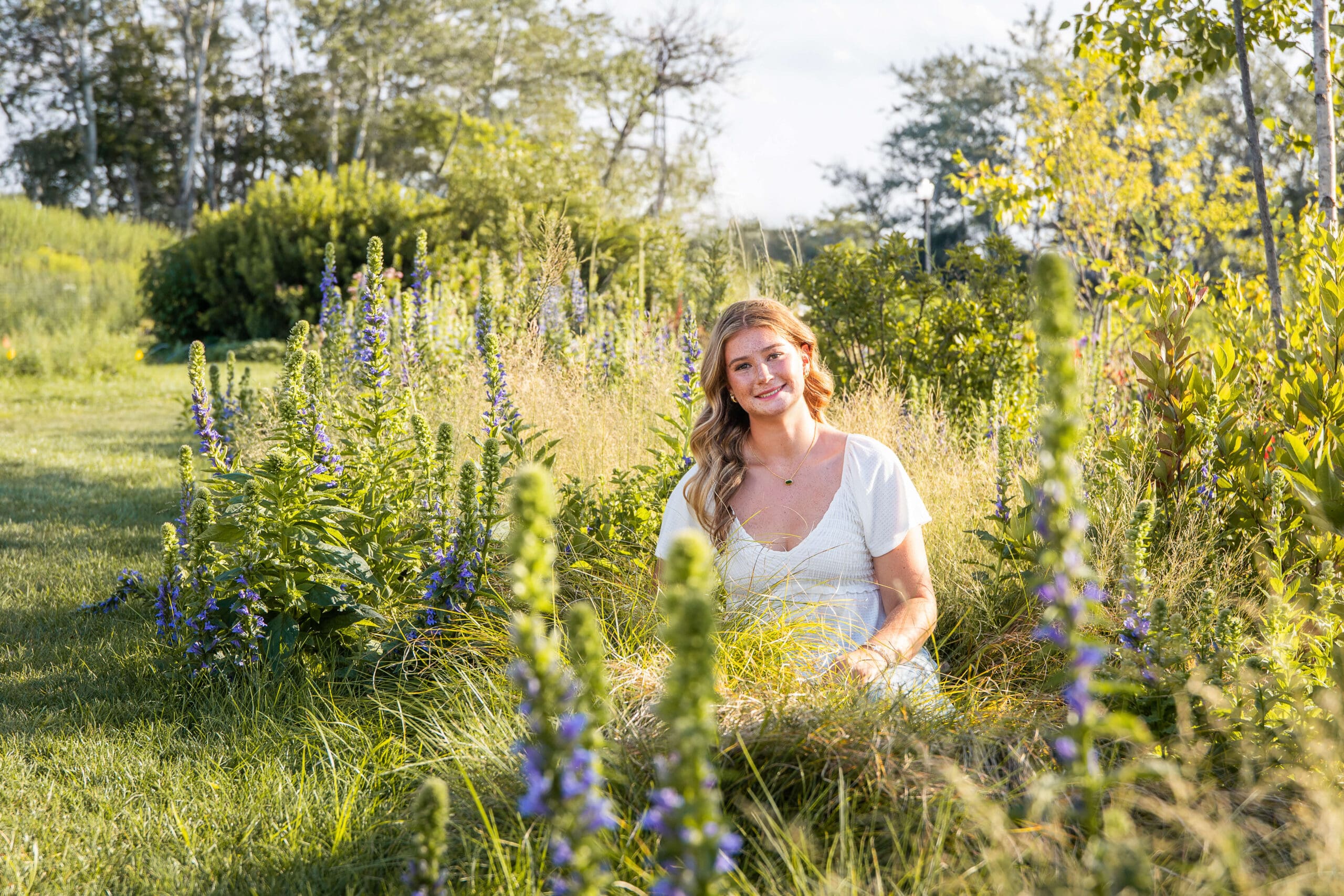 A high school senior in a white dress sits in a field of blue wildflowers smiling at sunset after visiting hair salons in metro detroit
