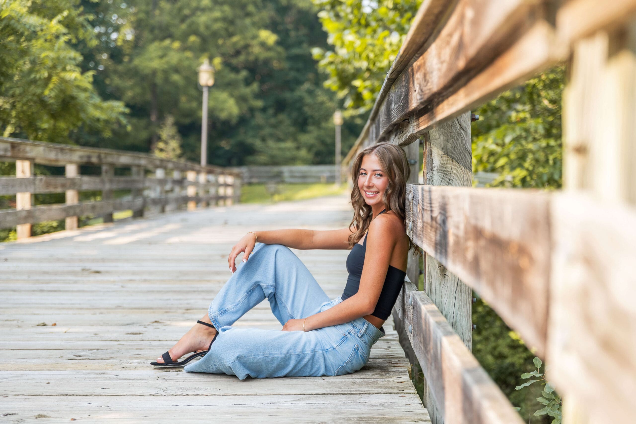 A happy high school senior in a black blouse and jeans sits on a boardwalk leaning on the railing after enjoying nail salons in metro detroit