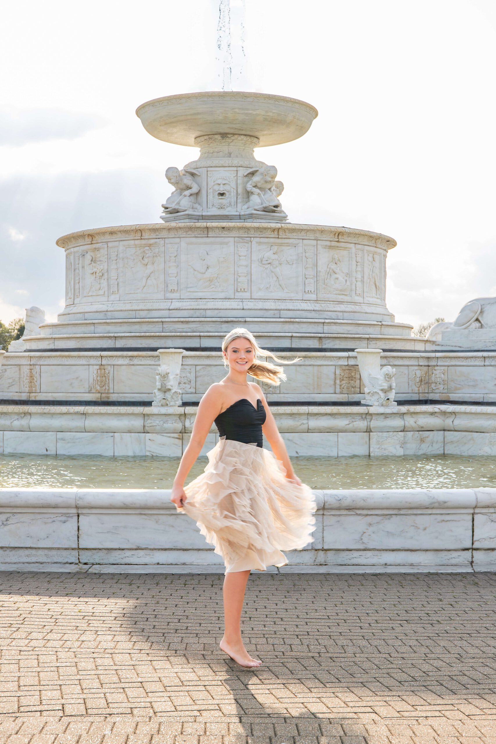 A high school senior dances in a tan and blue dress in a park in front of a large fountain at sunset after finding great prom dresses in detroit