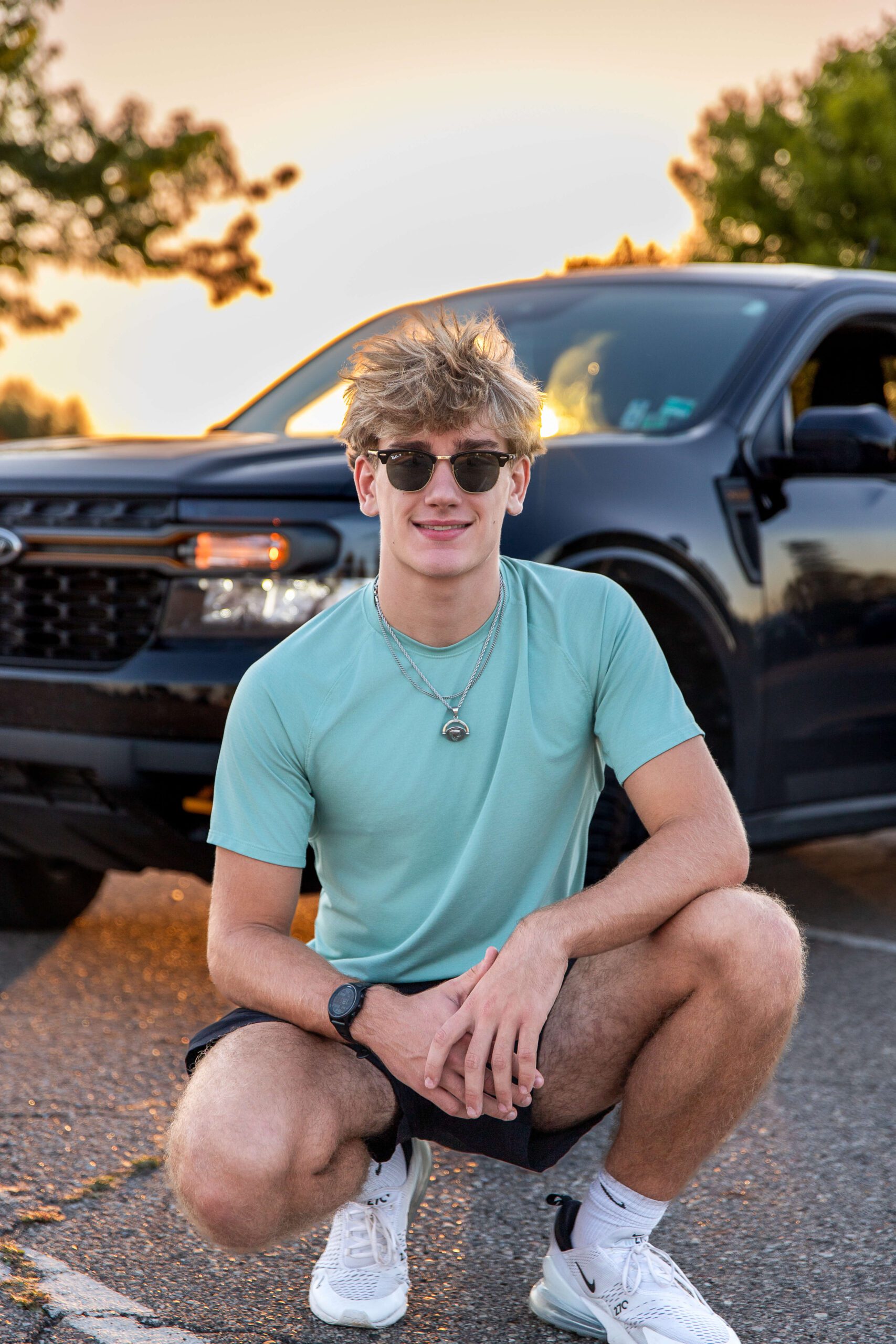 A smiling teenager in a teal shirt squats in front of a dark truck at sunset in a park with sunglasses after some sat prep in detroit