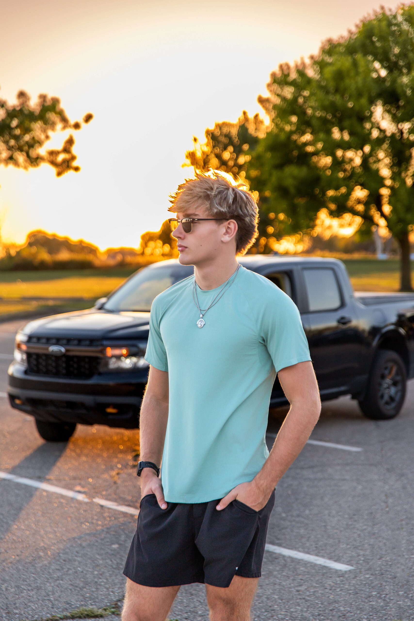 A high school senior stands in a teal shirt with hands in his pockets at sunset with sunglasses in a parking lot after some sat prep in detroit