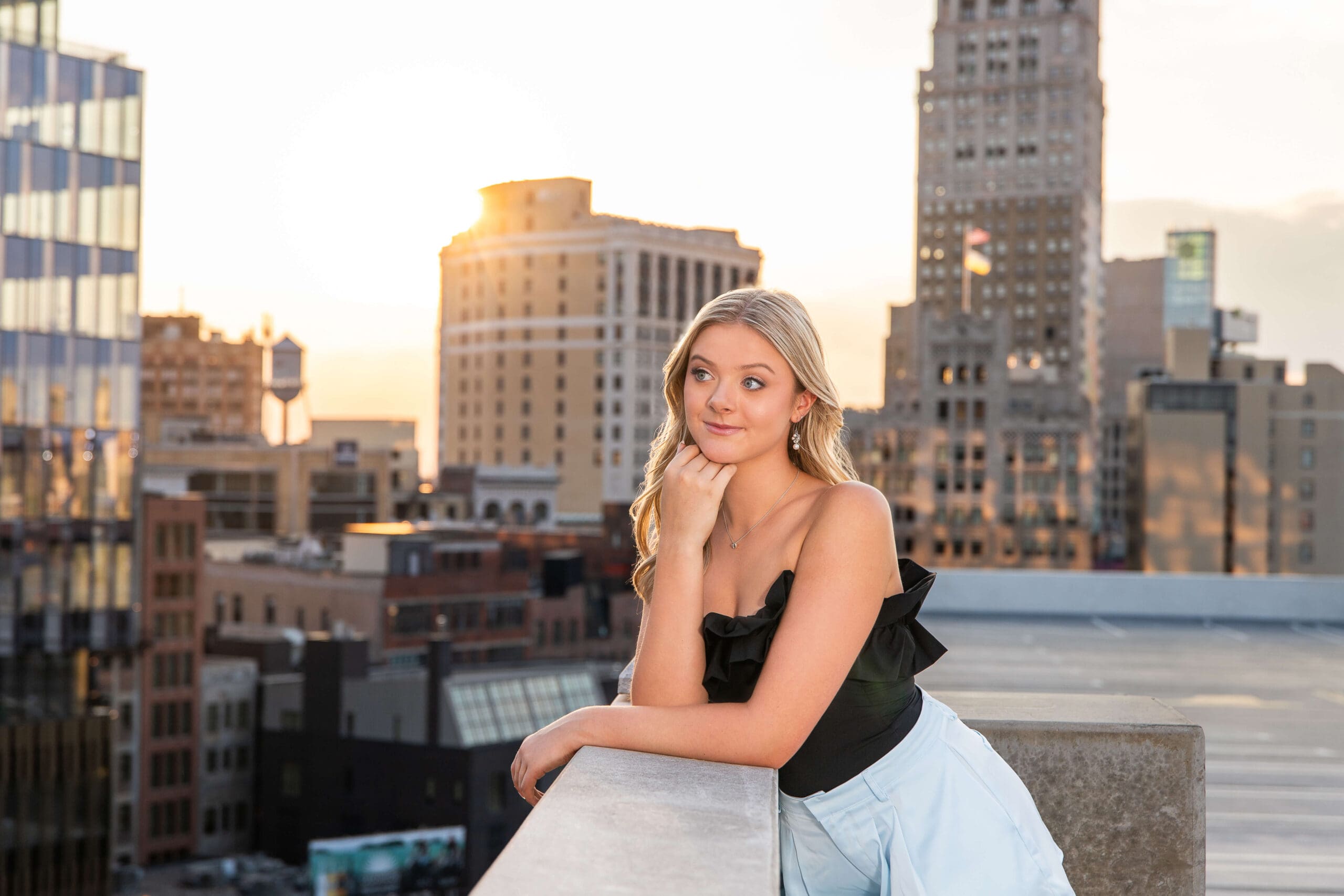 A high school senior in a white blouse leans on a parking garage wall overlooking the city at sunset after finding tanning salons in metro detroit
