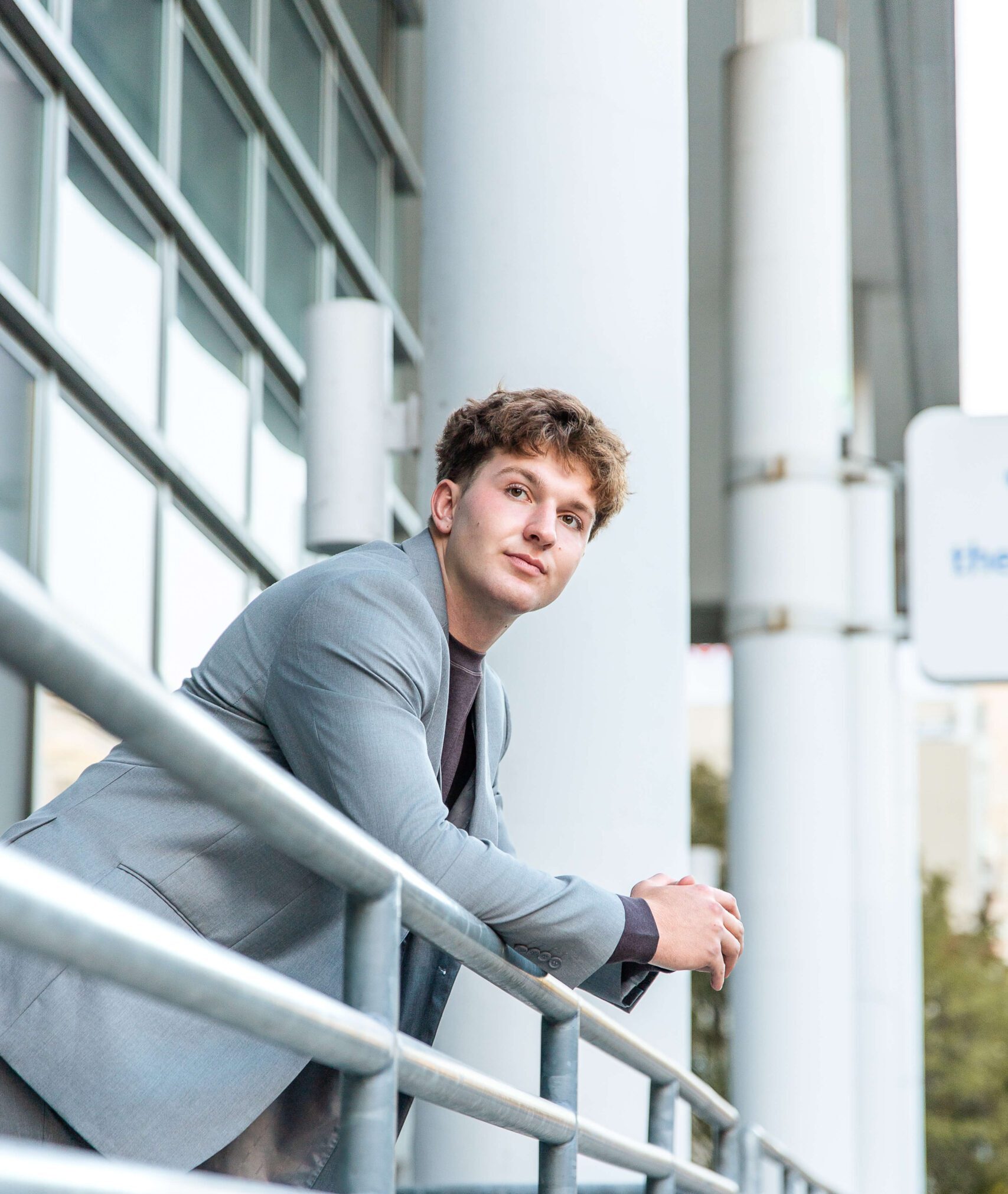 A high school grad in a grey suit leans on a metal railing of a large building at sunset