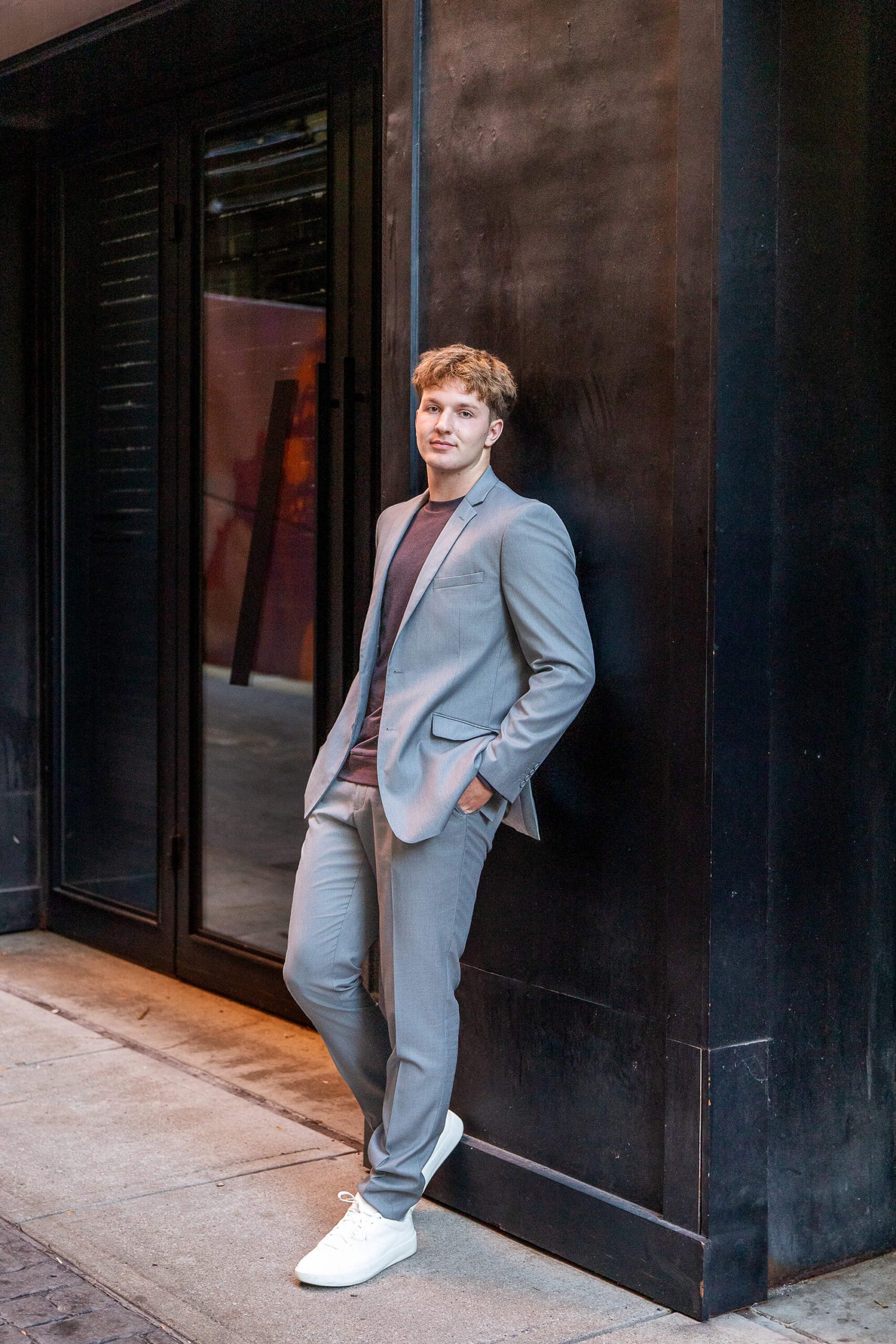 A high school senior leans on a black wall with hands in pockets in a grey suit after enjoying tux rental in detroit