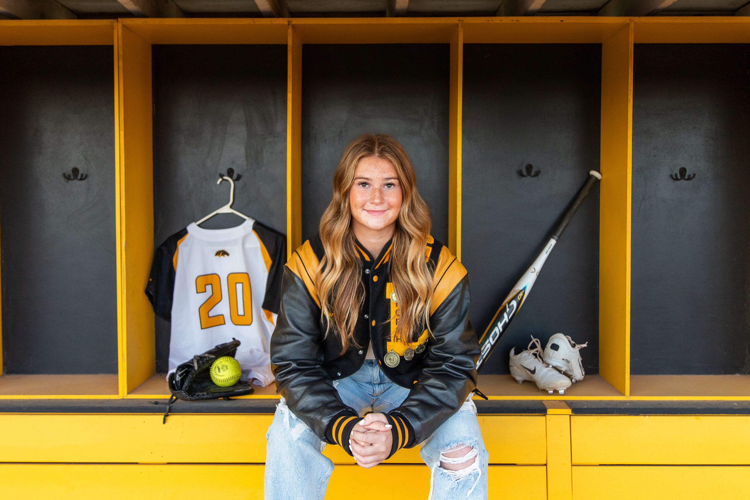 A smiling high school senior sits in a letterman jacket and jeans on a dugout bench with softball gear