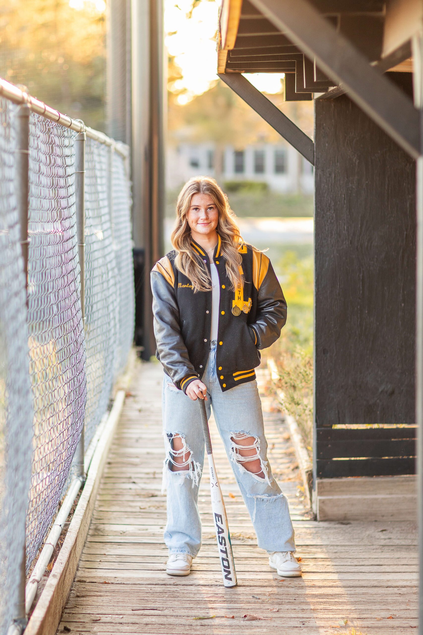 A high school senior softball player leans on a bat in a letterman jacket while standing in wooden dugout at sunset after finding volunteer opportunities in metro detroit
