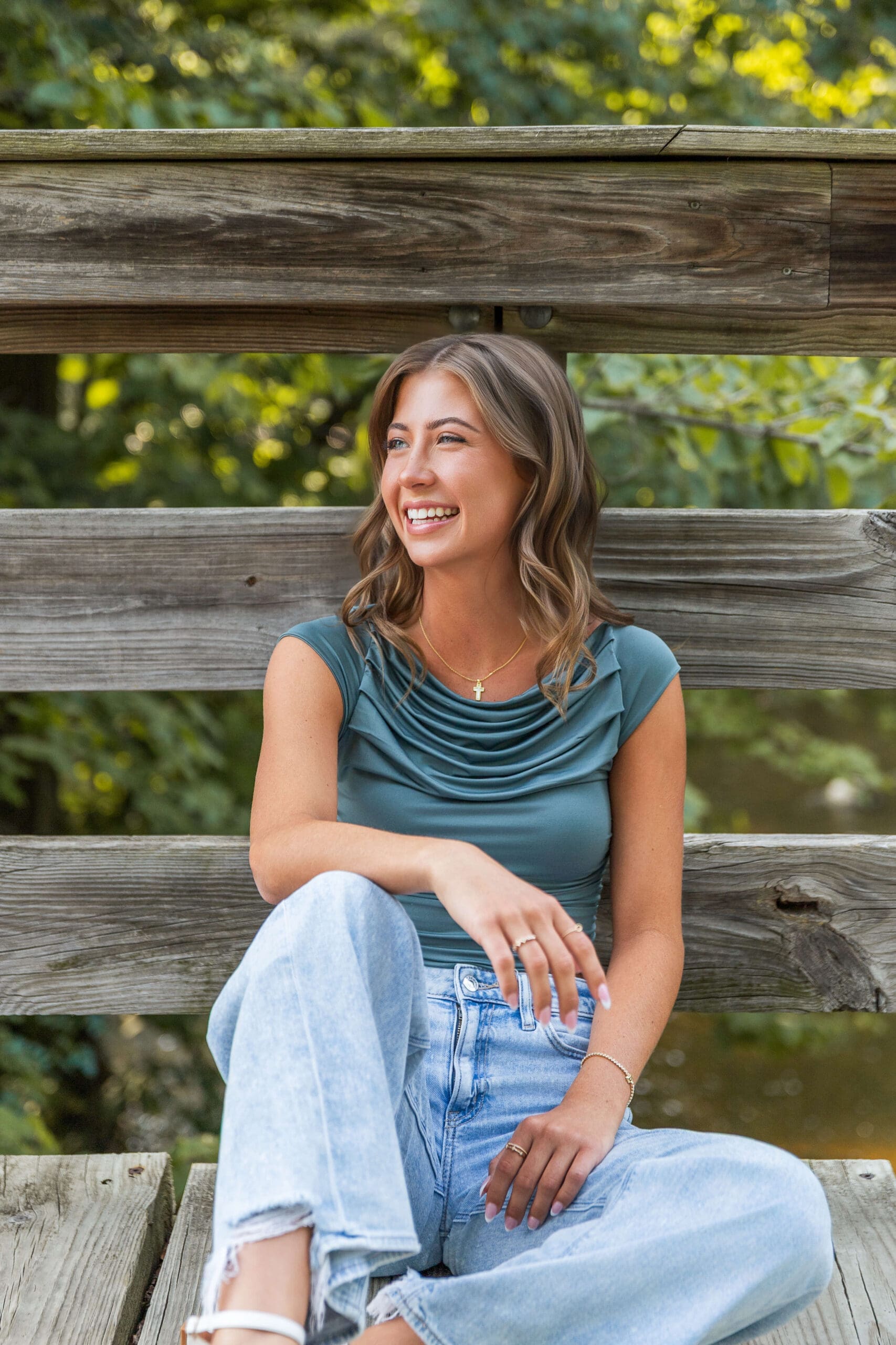 A laughing high school senior in a blue blouse and jeans sits on a wooden boardwalk after exploring colleges in michigan
