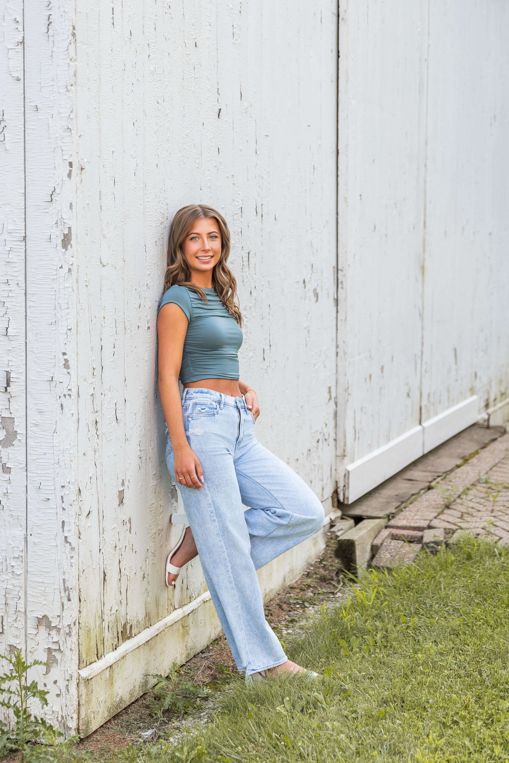 A high school senior in a blue blouse leans on a rustic white wall smiling after visiting colleges in michigan