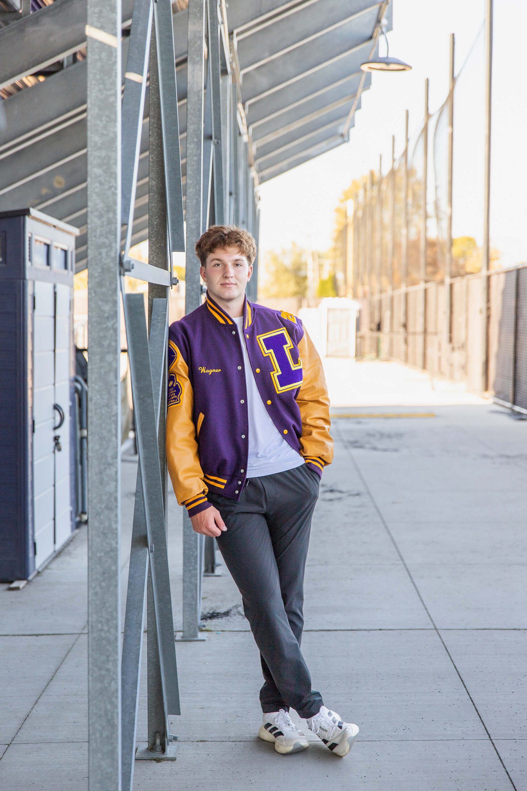 A high school senior leans under the bleachers on a support beam with a hand in his pocket in a purple letterman jacket