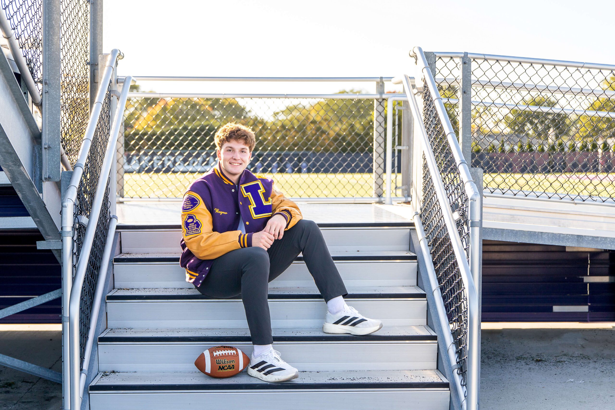 A teenager in a purple letterman jacket sits on stairs to the bleachers smiling at sunset after exploring colleges near detroit