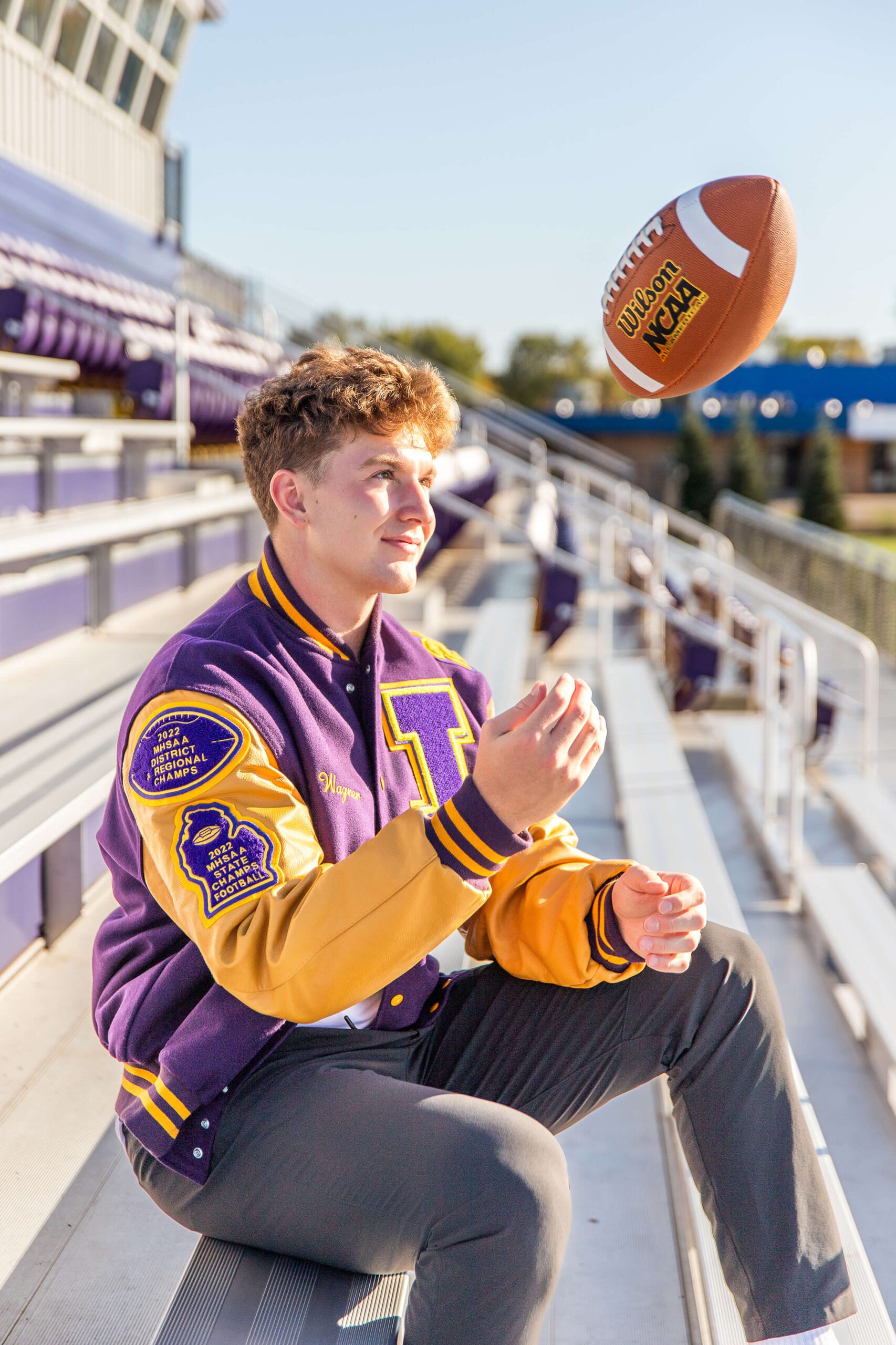 A happy high schooler in a purple letterman jacket suits in the stands tossing a football after visiting colleges near detroit