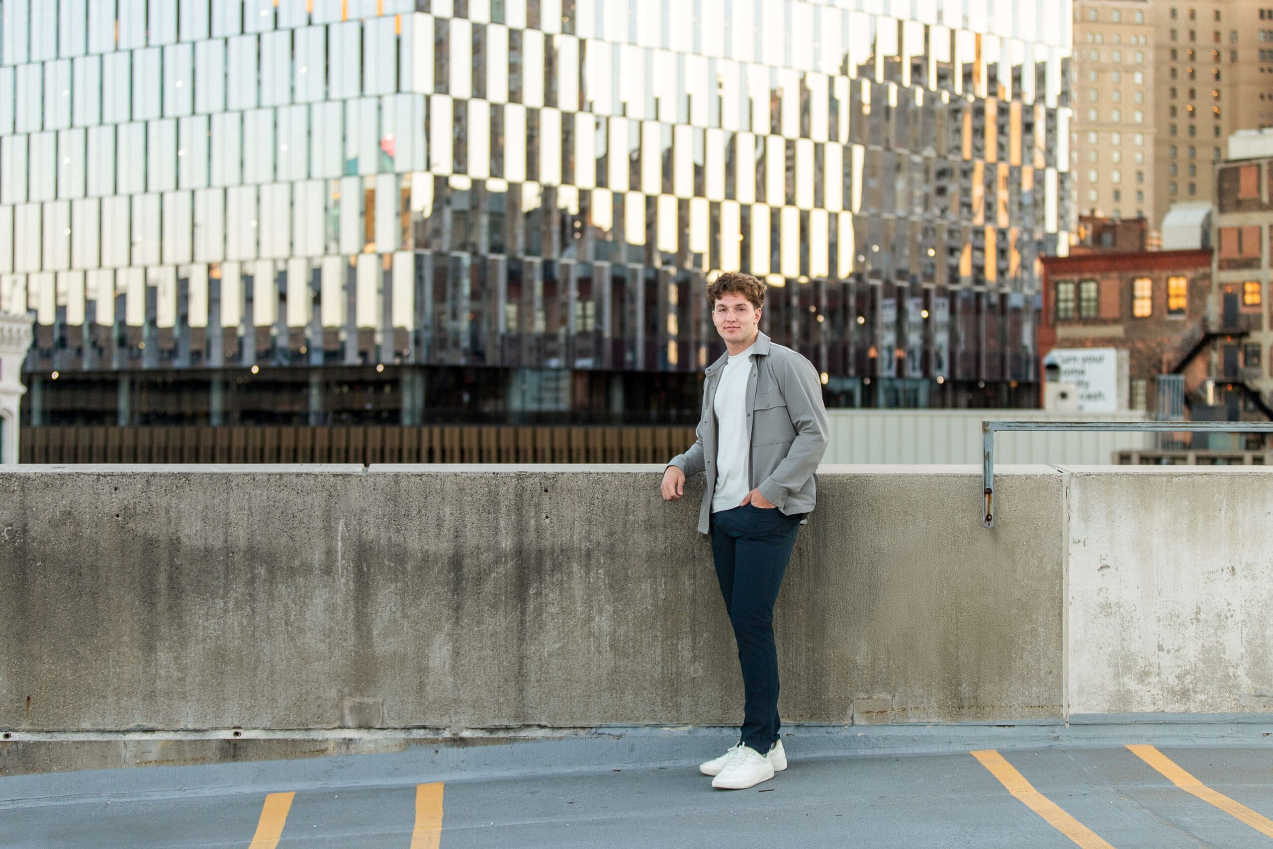 A man leans on the railing of a parking garage roof in a grey shirt and blue pants in downtown