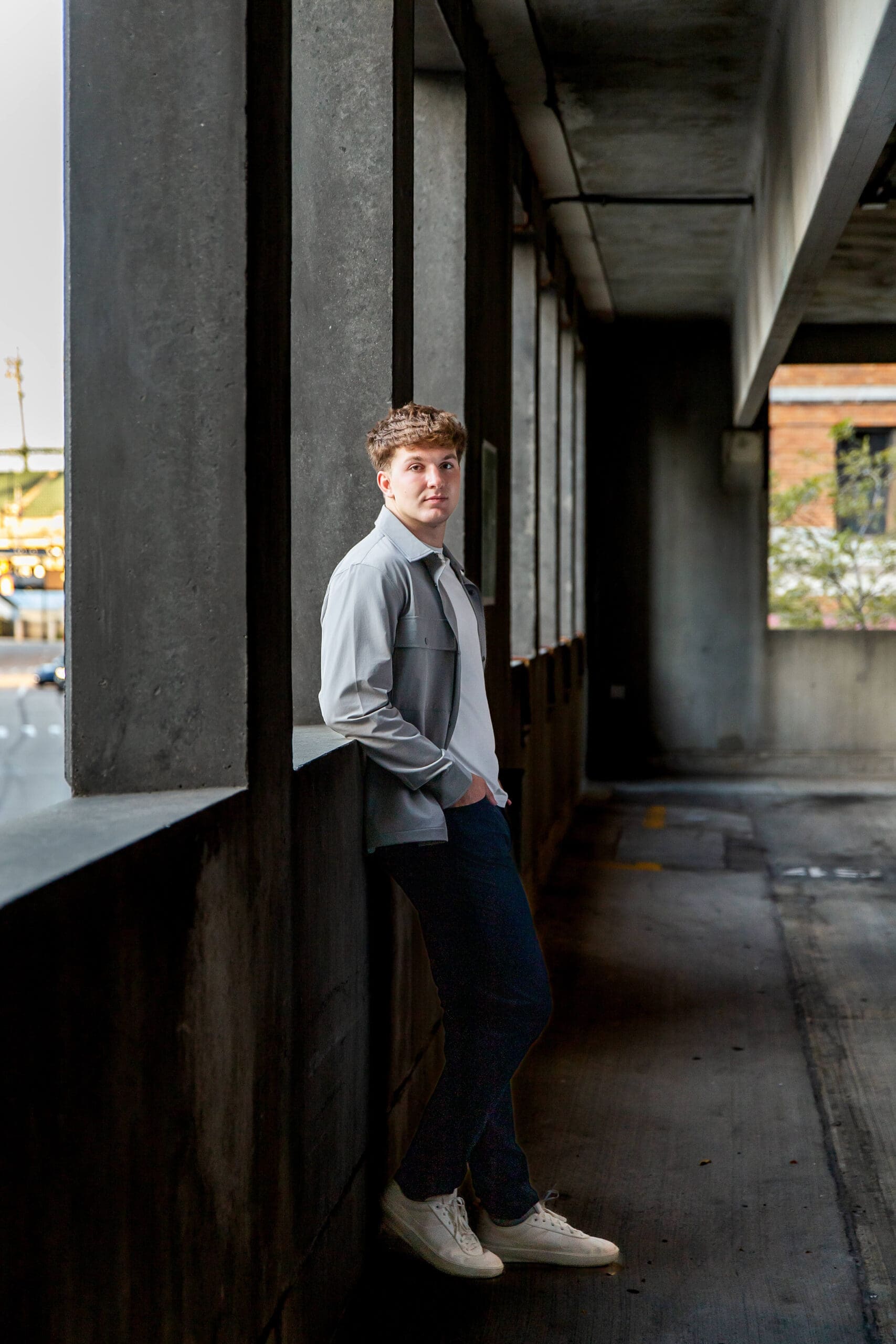 A high school grad leans against a parking garage wall in a grey shirt and jeans after visiting community colleges in detroit