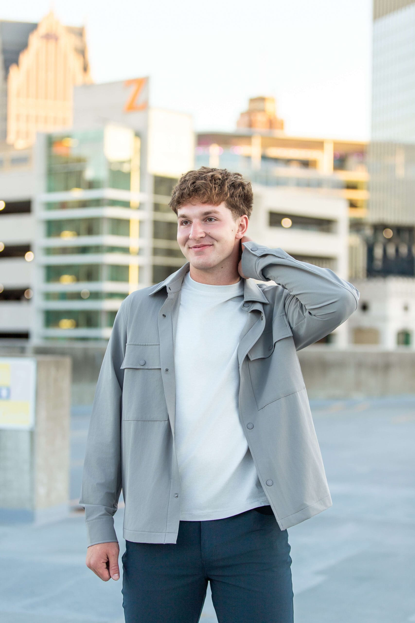 A high school senior in a grey shirt stands on a parking garage roof smiling at sunset after exploring community colleges in detroit