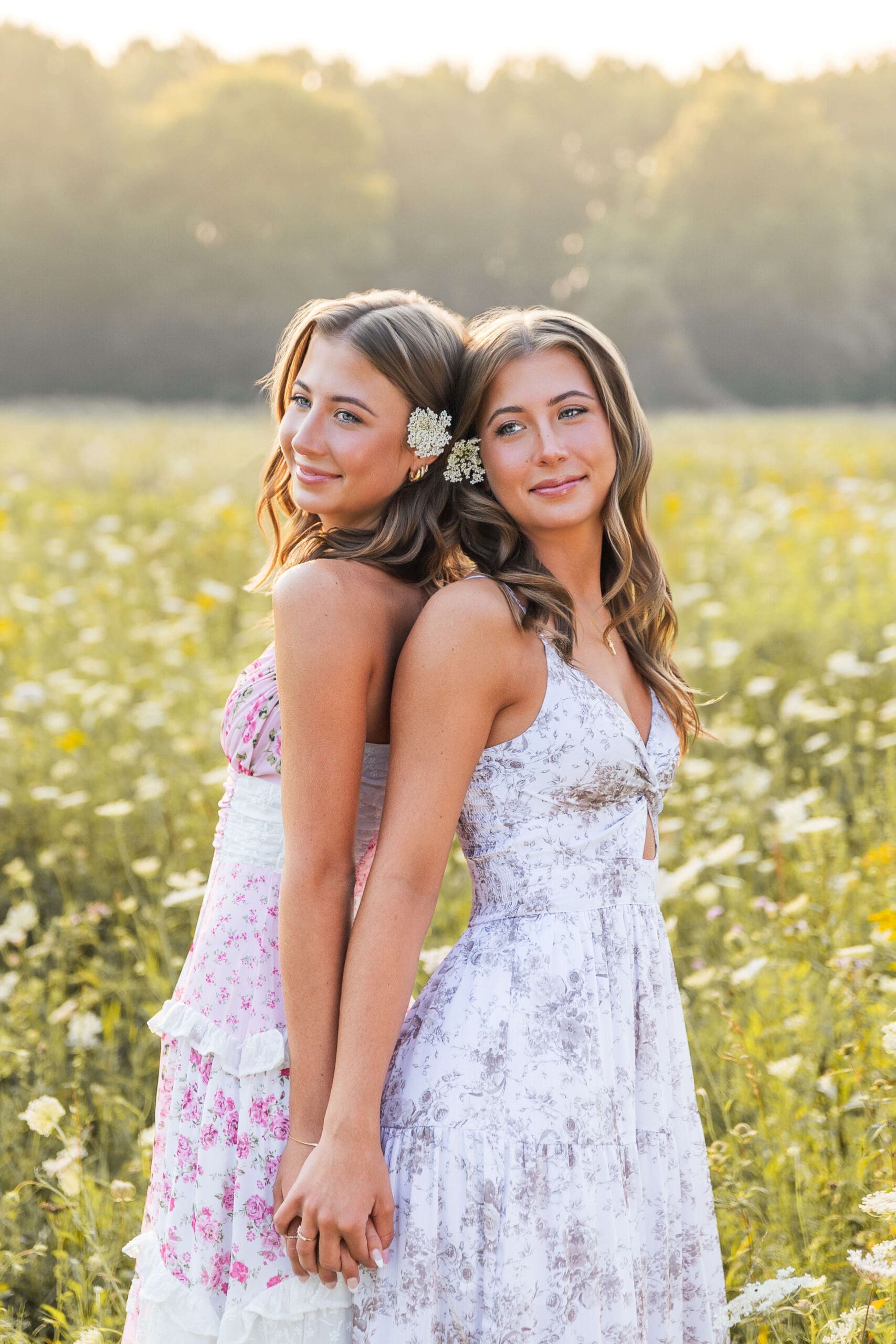 Twin high school senior sisters lean on each other in a field at sunset in floral print dresses