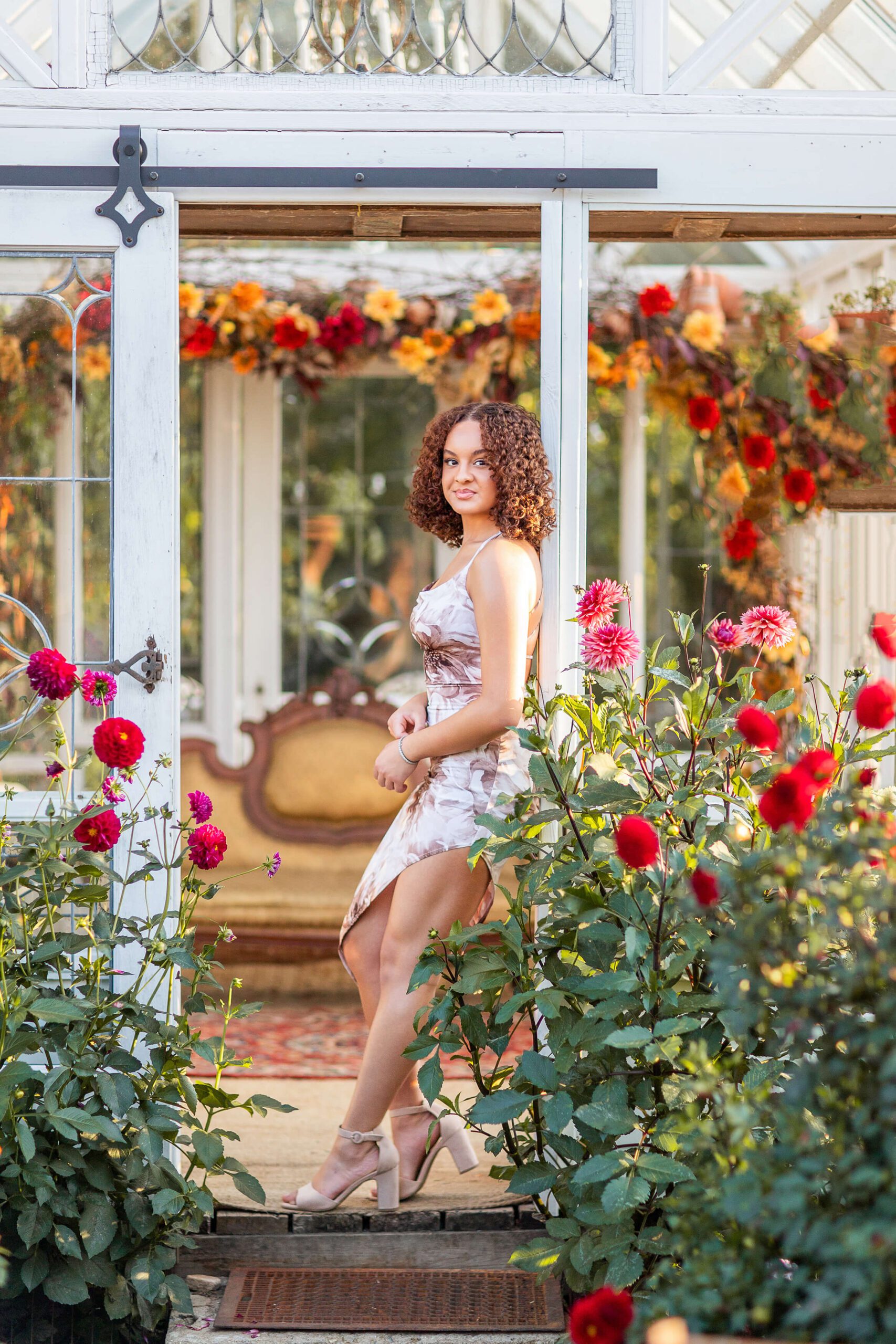 A high school senior in a floral print gown leans in a greenhouse entry surrounded by red roses after visiting hair salons in washington mi for her curls