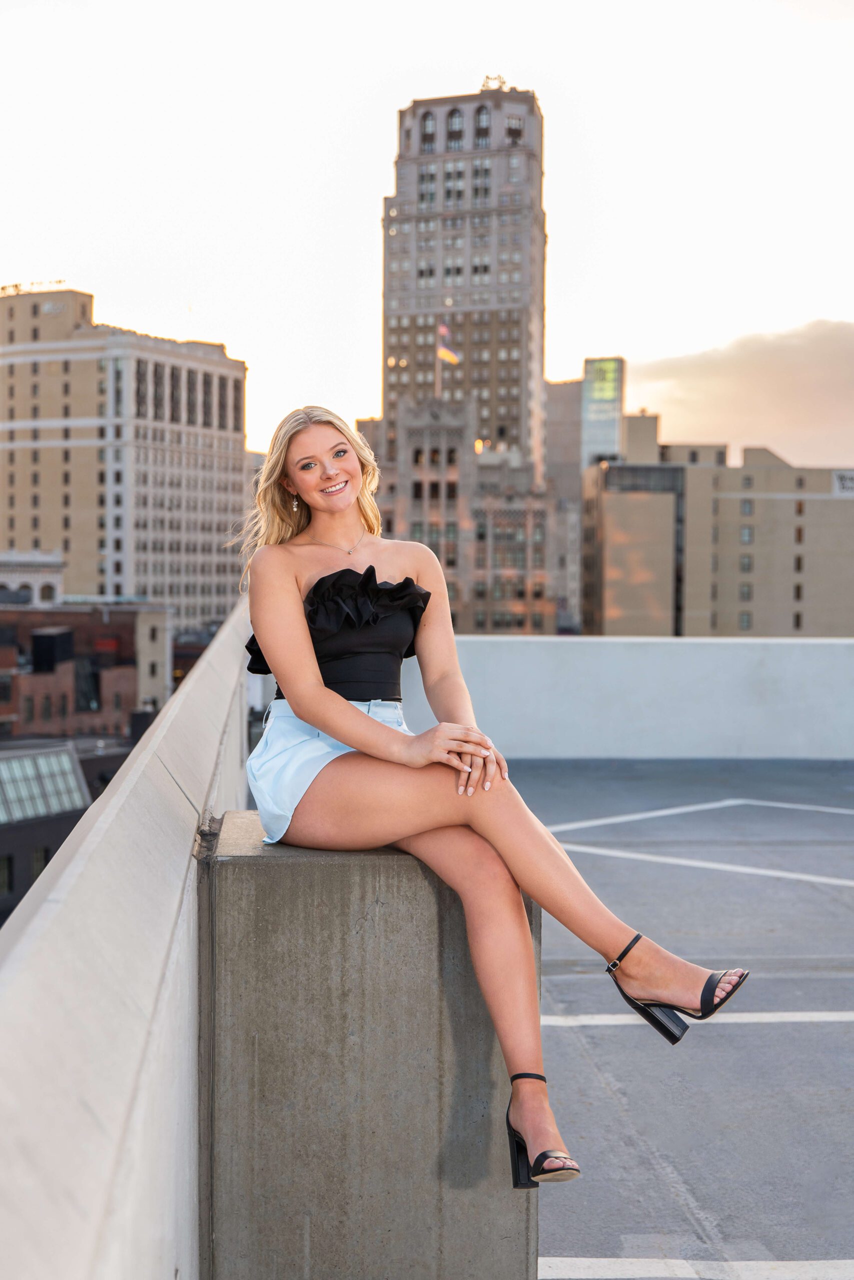A smiling blonde woman sits on a parking garage roof at sunset in downtown in a black blouse and short skirt