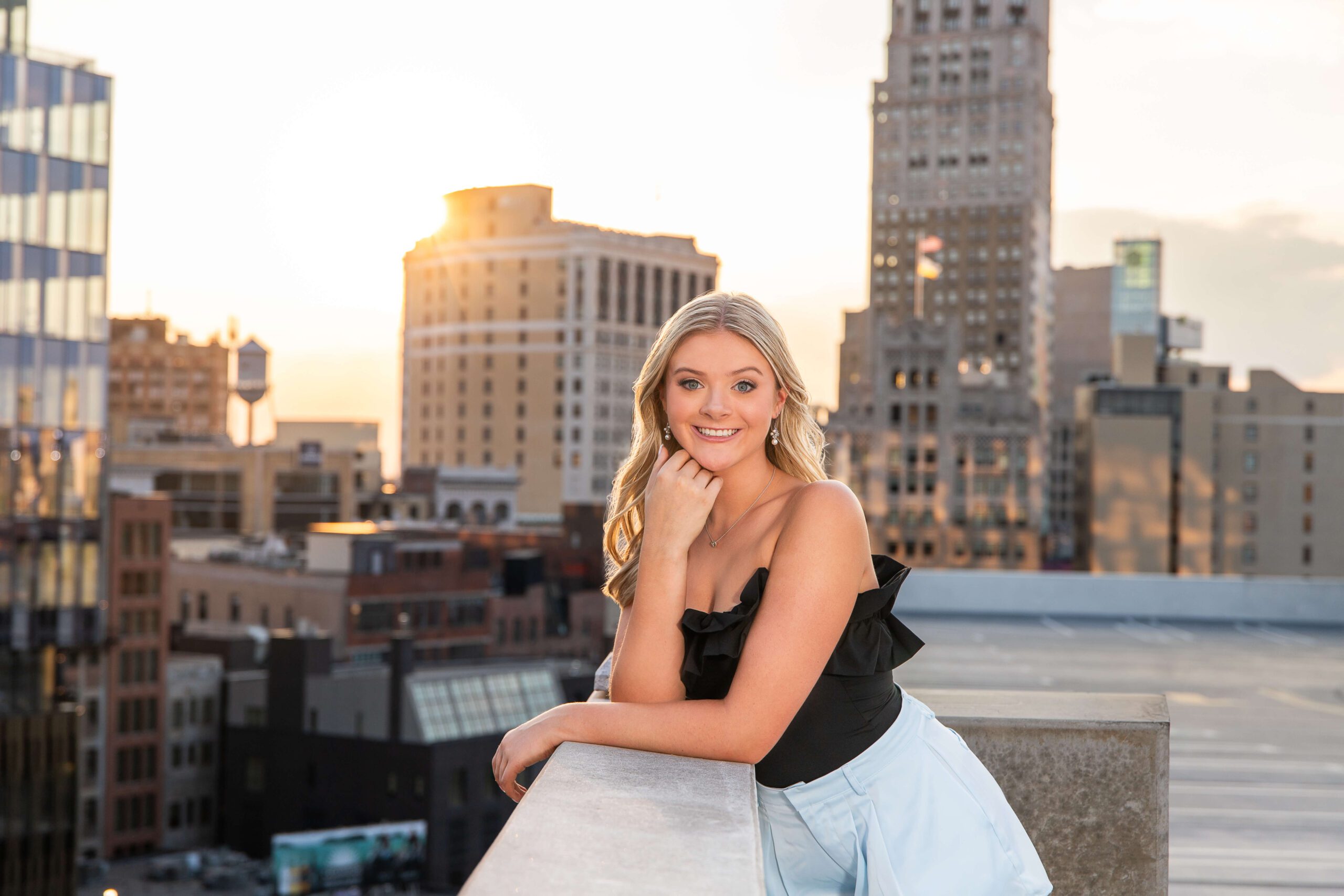 A blonde high school senior in a black blouse and blue skirt leans on a parking garage wall at sunset in downtown after finding nail salons in washington mi