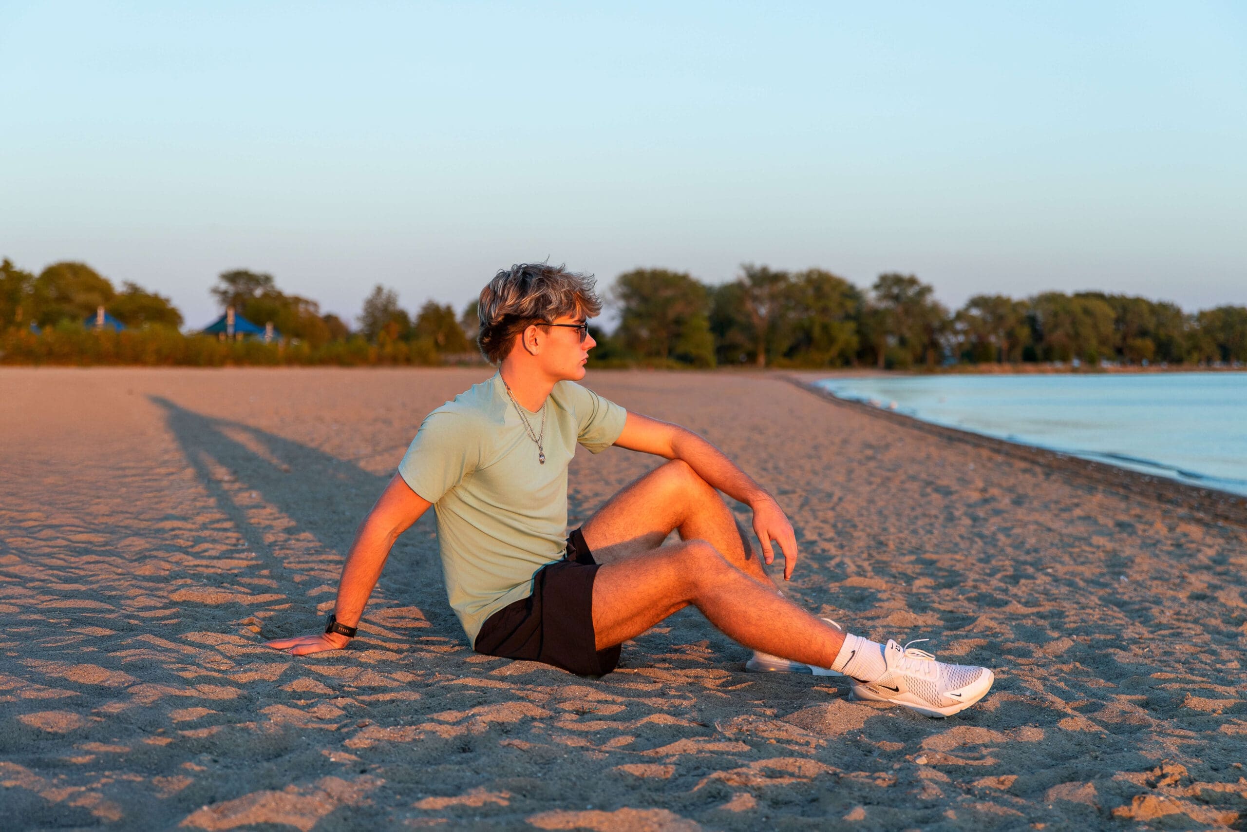 A high school senior sits on the beach in a green shirt and sunglasses at sunset during one of the things to do with teens in detroit